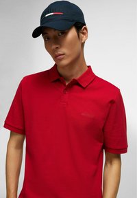 Rode polo shirt met een kraag en twee knopen, voorzien van subtiele "HECHTER" borduring. Draag het met een marineblauwe pet met logo en streepaccenten.