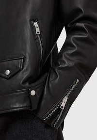 Giacca in pelle nera con zip metalliche, bottoni a pressione e una finitura leggermente testurizzata. Include tasche sul petto e sui lati.