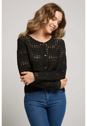 Femme souriante, portant un cardigan noir en crochet boutonné avec des motifs carrés et un jean bleu, bras croisés devant elle.