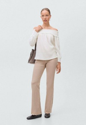 Vit avskuren blus med ribbad textur; ljusbeiga utsvängda byxor; modellen håller en brun väska och har på sig svarta skor med dekorationer.