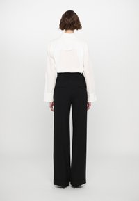 Camicia bianca a maniche lunghe con dettaglio sulla schiena, abbinata a pantaloni neri a gamba larga con due tasche posteriori e dettagli sul fondo.