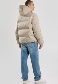 Giubbotto puffer beige con cappuccio, caratterizzato da un design trapuntato, indossato sopra jeans azzurri chiari e sneaker bianche, visto da dietro.