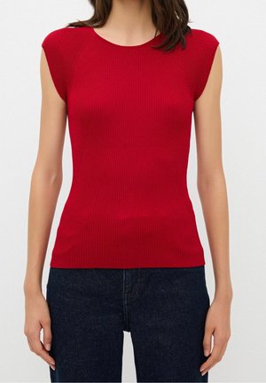 Ärmelloses rotes, geripptes Strickoberteil mit rundem Ausschnitt, getragen zu dunkelblauen Jeans an einer stehenden Person.