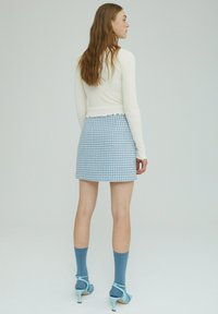 Weißes langärmeliges Oberteil mit gewelltem Saum, kombiniert mit einem blau karierten Miniröcke. Blaue knielange Socken und hellblaue Absatzschuhe runden das Outfit ab.