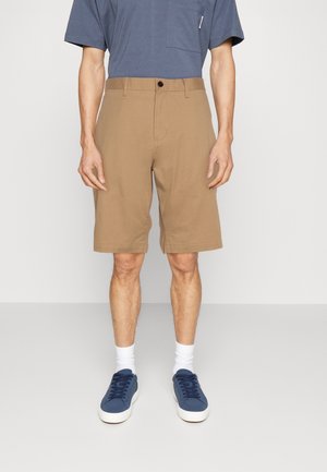 Homme portant un short beige jusqu'aux genoux, des baskets bleu marine, des chaussettes blanches et une chemise bleue à manches courtes, debout devant un fond uni.
