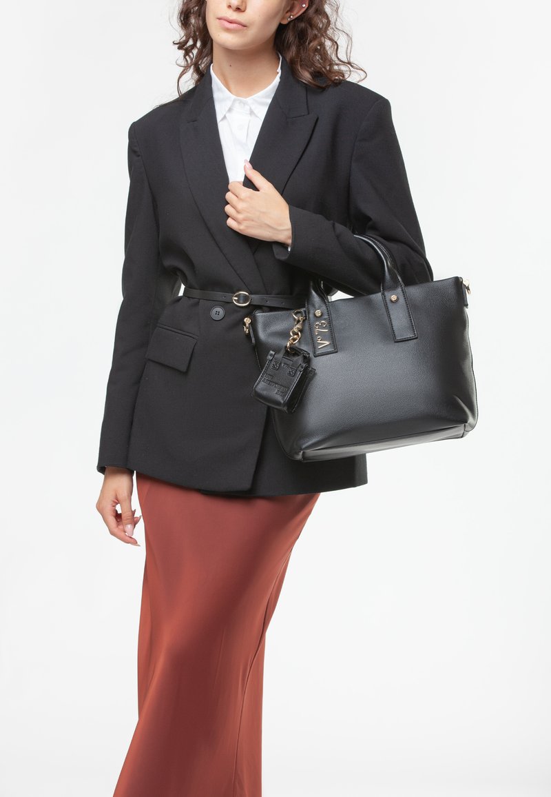Bolso de mano de cuero negro con asas dobles, textura suave, herrajes dorados y un pequeño charm adjunto. Lleva un blazer negro entallado y una falda marrón.