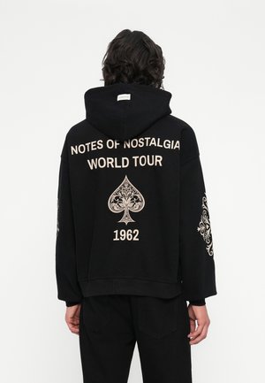 Črna kapuco majica s prevelikim krojem. Ima belo natisnjeno besedilo "NOTES OF NOSTALGIA WORLD TOUR 1962" in dekorativne grafike na rokavih.