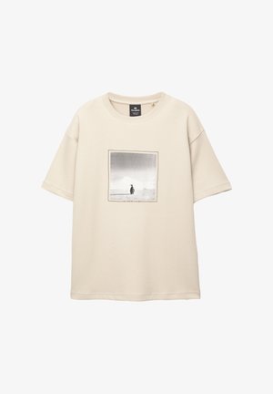 Beige Baumwoll-T-Shirt mit kurzen Ärmeln, das einen rechteckigen monochromen Druck einer Person vor einer Landschaft zeigt. Text unter dem Druck enthalten.