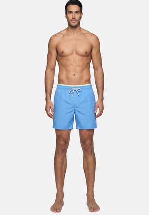 Gespierde man staat op blote voeten, draagt lichtblauwe zwemshorts met witte trekkoorden tegen een effen achtergrond.