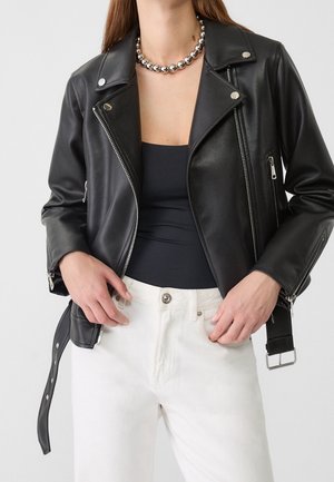 Femme portant une veste en cuir noire de motard, un haut noir, un pantalon blanc et un collier argenté à perles, mains près des poches du pantalon.
