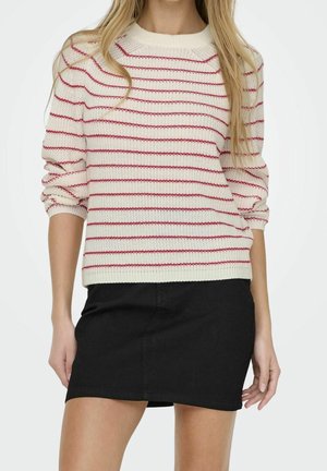 Femme portant un pull blanc avec de fines rayures rouges horizontales et une mini-jupe noire, debout les mains détendues sur les côtés.