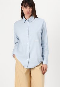 Pale blue marškiniai su mygtukais, pagaminti iš medvilnės, su laisvu kirpimu, klasikiniu apykaklės dizainu ir balta siuvinėta logotipu ant krūtinės.