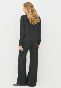 Sort bluse med lange ærmer og rund halsudskæring, parret med sorte jeans med vide ben, der har gule stikninger og baglommer.