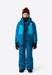 Costum de schi în albastru vibrant, care include o jachetă impermeabilă cu design color-block, pantaloni salopetă izolați, mănuși și ochelari de schi.