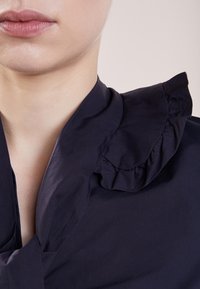 Blusa blu navy con colletto a volant, caratterizzata da una texture liscia e un design strutturato, primo piano del collo e della parte superiore del petto.