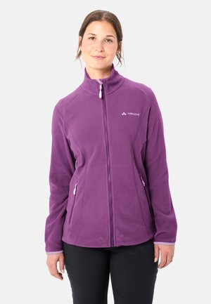 Frau trägt eine lila Fleecejacke mit Reißverschluss und hohem Kragen, steht vor einem schlichten hellen Hintergrund, die Hände entspannt an den Seiten.