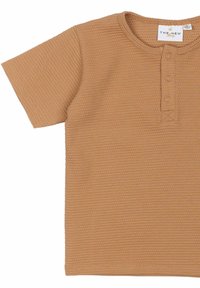 Krátký rukáv světle hnědého trička z texturované pleteniny ve vaflovém vzoru, s kulatým výstřihem a detailem tří knoflíků na léze.
