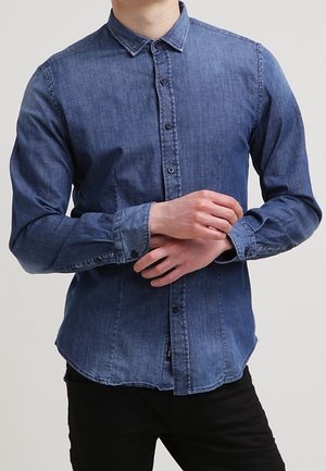 Mann trägt ein langärmeliges blaues Denimhemd, das zugeknöpft ist, und justiert eine Manschette, steht vor einem schlichten hellen Hintergrund.