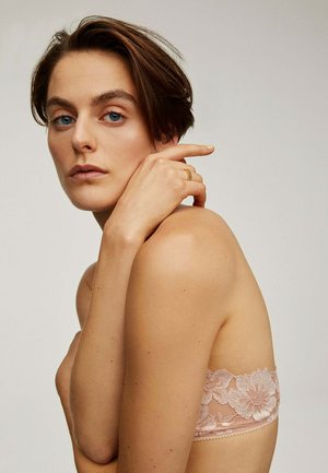 Bralette de encaje en rosa suave, con patrones florales. La modelo posa con la mano en el hombro, mostrando la textura y los delicados detalles de diseño.