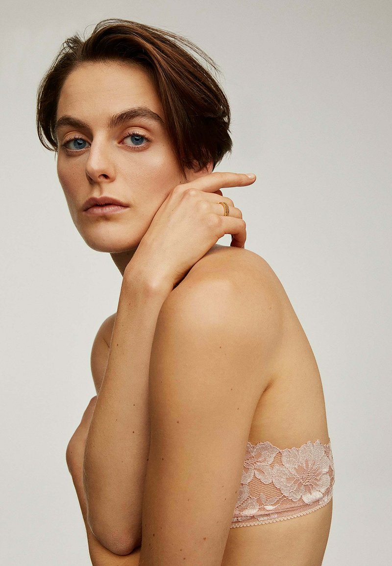Bralette de encaje en rosa suave, con patrones florales. La modelo posa con la mano en el hombro, mostrando la textura y los delicados detalles de diseño.