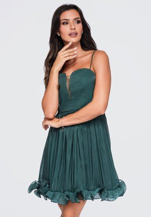 Mujer con cabello largo y oscuro vistiendo un vestido plisado sin mangas de color verde oscuro con dobladillo con volantes, posando con una mano en la barbilla y la otra en la cintura.
