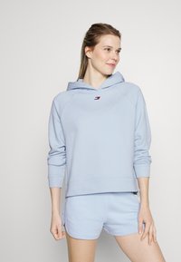 Jasnoniebieska bluza z kapturem wykonana z miękkiego materiału, z kieszenią z przodu, ściągaczami przy rękawach i małym logo. W połączeniu z dopasowanymi szortami dla uzyskania swobodnego wyglądu.