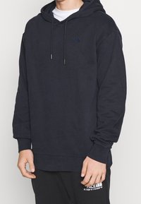 Námořnicky modrá mikina s kapucí z bavlněné směsi, s přední kapsou kangaroo, stahovací šňůrkou na kapuci a malým logem na hrudi. Měkká textura.