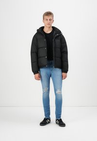 Chaqueta de plumas negra con grandes costuras en forma de cuadrado, bolsillos frontales y una capucha, combinada con jeans claros rasgados y zapatillas negras.