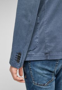 Niebieska bawełniana kurtka z jedną kieszenią i szczegółem czterech guzików na mankietach, zestawiona z niebieskimi dżinsami z typowymi akcentami ściegowymi.