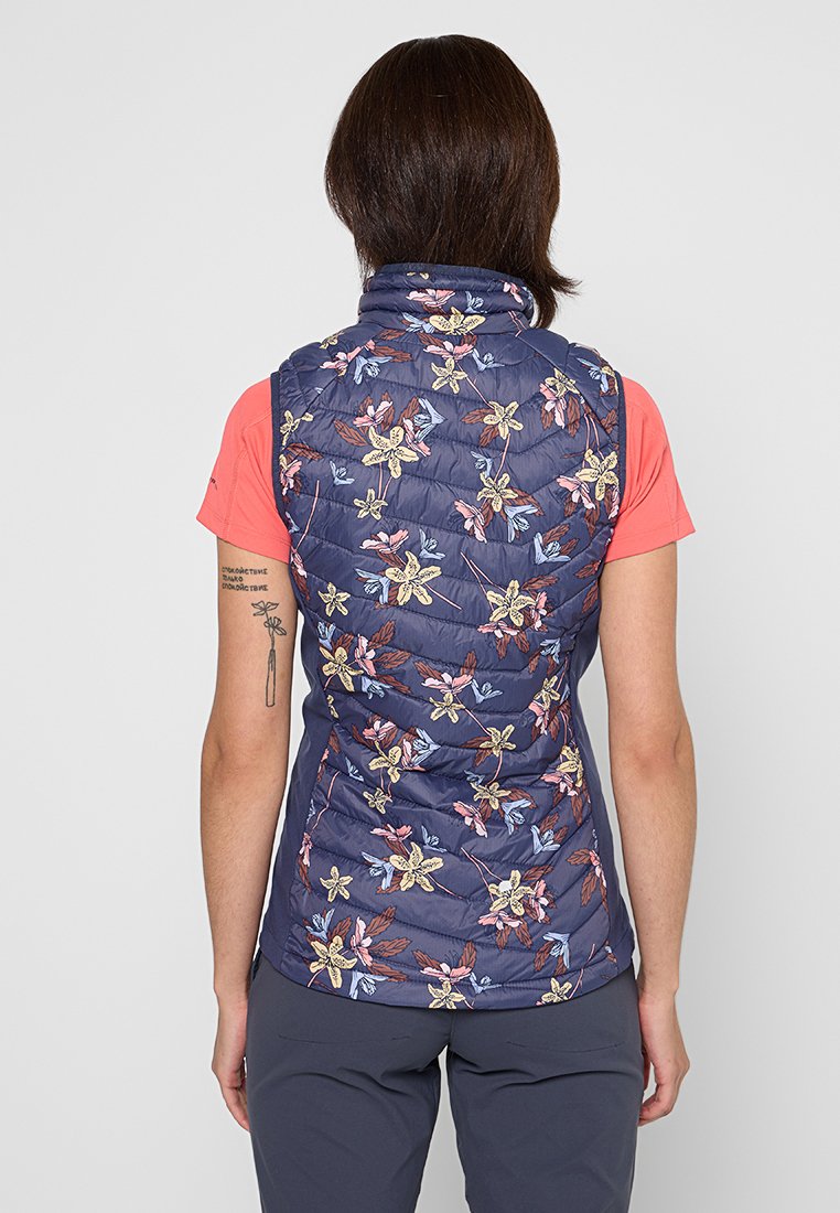 Chaleco floral en navy con textura acolchada, que presenta patrones de flores multicolores. Llevado sobre una camisa rosa, con un cuello alto y diseño sin mangas.