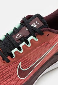 Baskets Nike avec tige bordeaux et rouge, lacets noirs, accents vert menthe, maille texturée, et semelle blanche. Marque notable sur la languette.