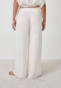 Pantalones ligeros, de pierna ancha, en color crema con una cinturilla elástica y una textura suave, mostrados desde la parte de atrás sobre una superficie texturizada.