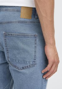 Svetlo modre kavbojke iz denima z zadnjim žepom in tan oznako iz usnja nad njim. Tkanina ima gladko teksturo z rahlim bledenjem.