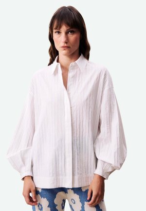 Vrouw draagt een loszittend wit gestreept overhemd met knopen, met volumineuze mouwen en een blauwe broek met grote witte bloemenpatronen.