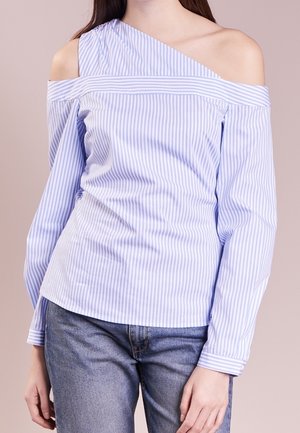 Vrouw die een blauw-wit gestreepte off-shoulder blouse draagt, gecombineerd met een blauwe spijkerbroek, staand tegen een neutrale achtergrond.