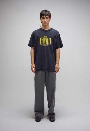 Ung person iført løstsiddende mørkegrå bukser, sorte sko og en sort T-shirt med et gult katte-motiv og teksten "Former".