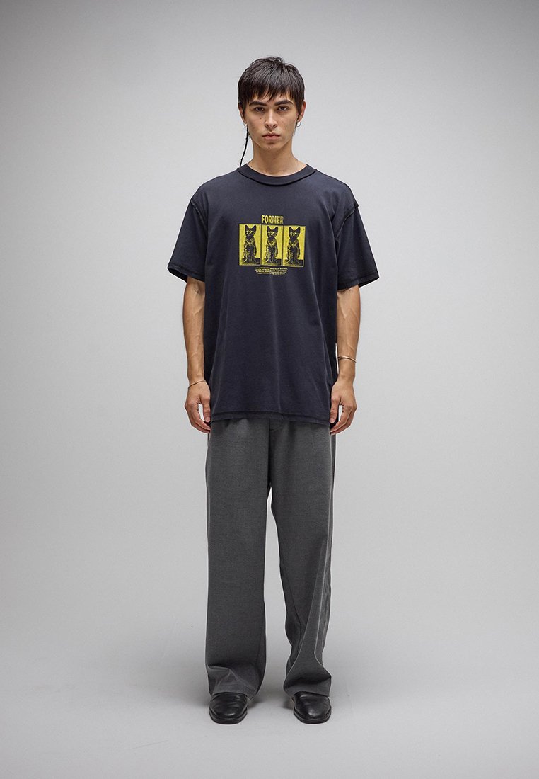 Νεαρός που στέκεται φορώντας φαρδιά σκούρα γκρι παντελόνια, μαύρα παπούτσια και μαύρο μπλουζάκι με κίτρινο γραφικό γάτας και το κείμενο «Former».