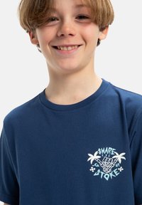 Tmavě modré bavlněné tričko s grafickým potiskem na levé straně hrudi, zobrazující palm trees a text "SDÍLEJTE NADŠENÍ" v světlé modré barvě.
