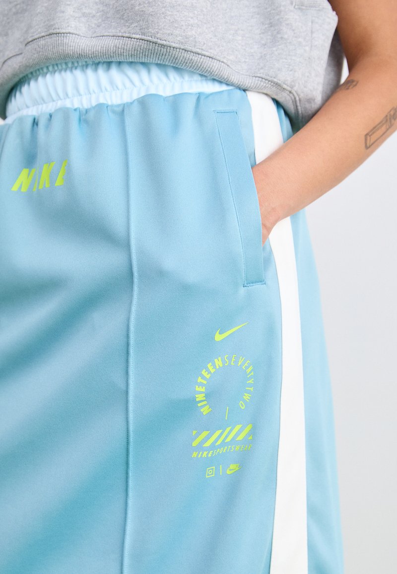 Pantaloni sportivi azzurri chiari con cintura elastica, strisce laterali bianche e dettagli grafici gialli sulla coscia sinistra. Marchio Nike visibile.