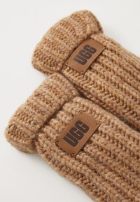 Gants en tricot marron avec une texture épaisse et une étiquette en cuir UGG sur chaque poignet. Des coutures renforcées le long des bords ajoutent de la durabilité.