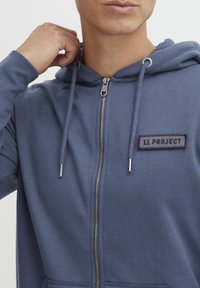 Felpa blu con zip in tessuto morbido, dotata di cordini regolabili, zip argentata e una patch nera con il logo "11 PROJECT" sul petto.
