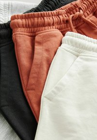 Kolme paria collegehousuja mustana, ruosteen värisenä ja off-white -värinä, joissa on joustovyötärö ja sivutaskut, esitelty teksturoidulla taustalla.