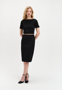 Czarna dopasowana sukienka z krótkimi rękawami, kwadratowym dekoltem i beżowym paskiem. Modelka nosi czarne lakierowane szpilki. Prosty i elegancki design.