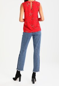 Blusa rossa senza maniche con motivo floreale, dettaglio a chiave sul retro, abbinata a jeans in denim blu e stivaletti neri con tacco alto.