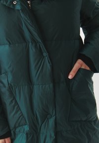 Zateplený zelený kabát s předním zapínáním na knoflíky, bočními kapsami a vysokým límcem. Hladký, jemný povrch s prošívanými segmenty.