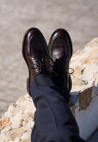 Zapatos de vestir de cuero marrón con una textura suave, punta redonda y cordones, colocados sobre una superficie de piedra, combinados con pantalones azul marino.