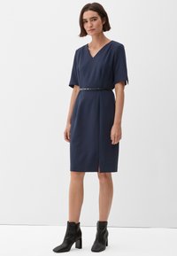 Navyblauwe jurk met korte mouwen, V-hals, ceintuurlijn en een split aan de voorkant. Gecombineerd met zwarte enkellaarsjes. Gladde stofafwerking.