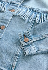 Hellblaues Jeansjacke mit Fransendetails an den Schultern, metallenen Druckknöpfen und Kontrastnähten. Weicher Stoff und lässiges Design.