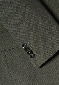 Gros plan sur le tissu d’une manche de costume texturée vert foncé avec quatre boutons noirs et une couture de poche visible.