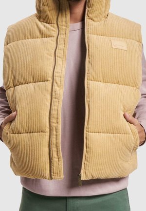 Beige kordfløjlsvest med lynlås, båret åben over en pink langærmet trøje med vaffelmønster og grønne bukser, hænderne i vestens lommer.